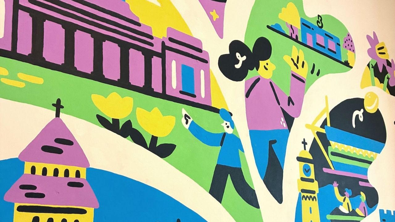 Colourful mural featuring abstract figures, buildings, and flowers in bright pink, blue, yellow, and green. The scene conveys a vibrant, playful tone.