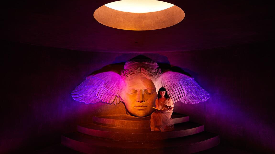 A serene woman sits on steps beside a large sculpted head with illuminated wings in a dimly lit room, creating a mystical and contemplative atmosphere.