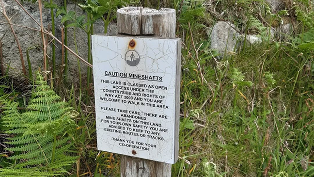 Sign on a wooden post in a grassy area warns of abandoned mineshafts. It advises caution and to stay on marked paths for safety.