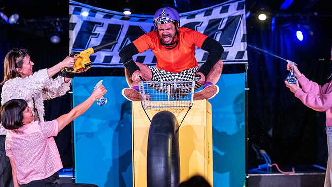 A man in a helmet, wearing a red shirt and checkered shorts, excitedly rides a makeshift vehicle with a large wheel. Two people on either side cheer him on with props. A "FINISH" banner hangs behind against a stage backdrop, creating an energetic and humorous atmosphere.