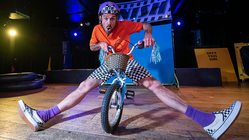 a man in an orange shirt and checkered shorts is spreading his legs towards the camera sat on a small blue bike.