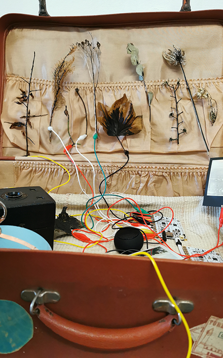 An open suitcase contains tangled wires connected to various electronics, including a camera. The setup appears chaotic, hinting at a DIY project.