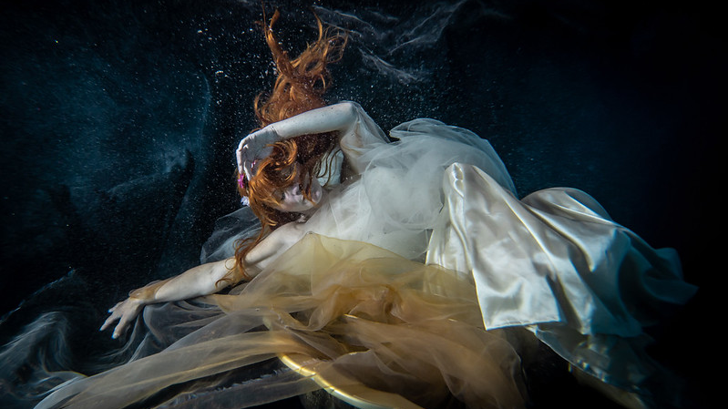 A woman with red hair floats gracefully underwater, wearing a flowing, translucent white gown creating an ethereal and serene atmosphere.
