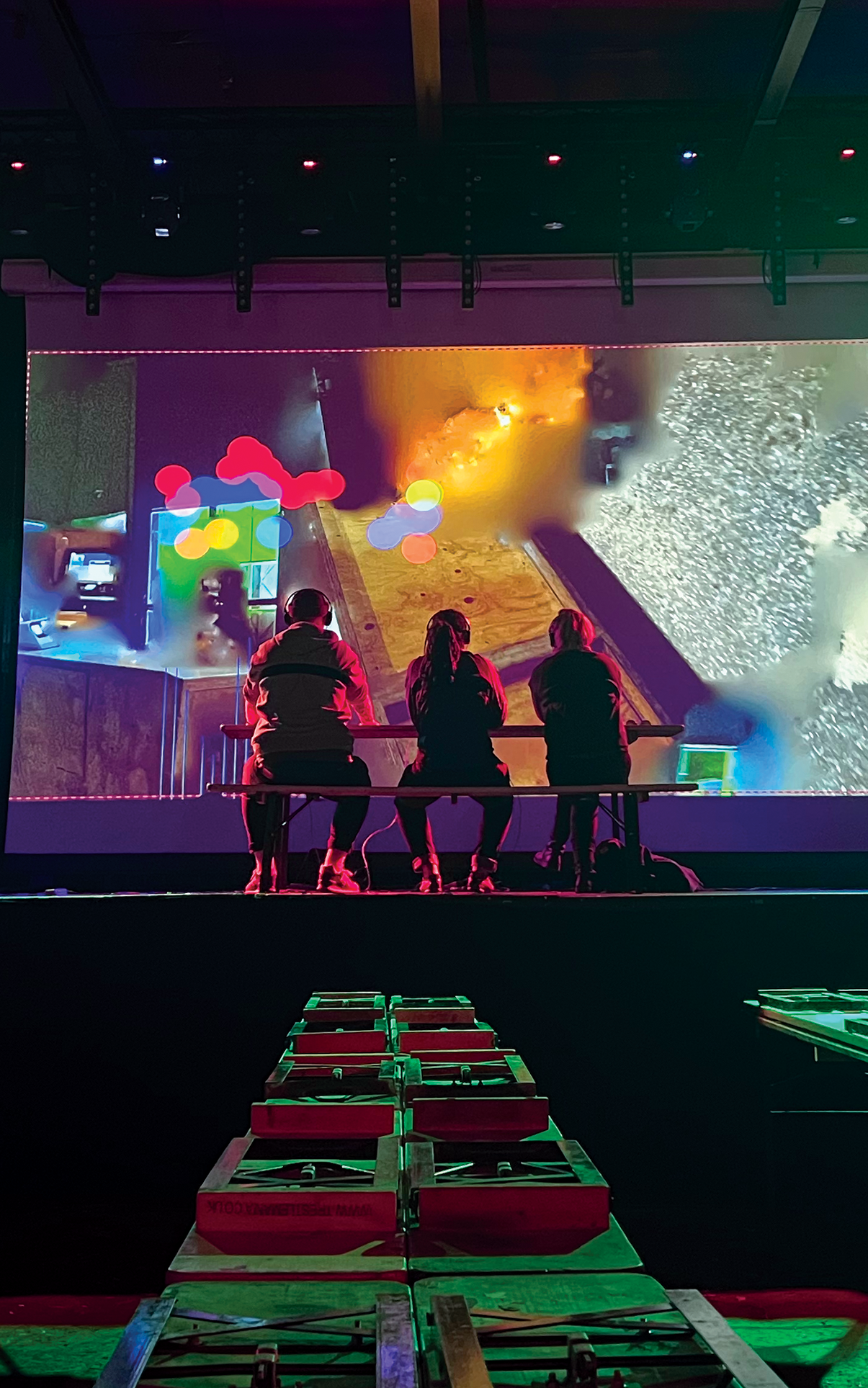 Three people sit on a stage facing a screen, there are objects on tables in the foreground below the stage, and the scene is lit in red and green.
