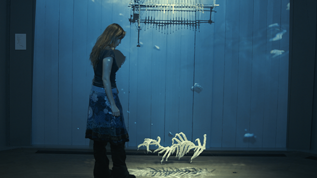 A woman gazes at a blue art installation resembling delicate white coral or sea life. The scene is softly lit, creating a contemplative and serene atmosphere.