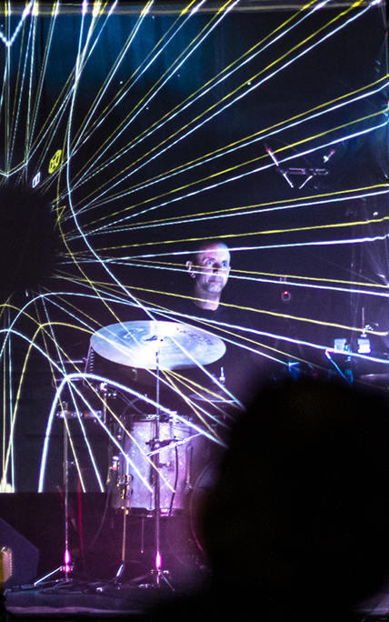 A drummer plays a set, surrounded by dynamic laser beams creating a vibrant, energetic atmosphere. The scene has a futuristic and immersive concert vibe.