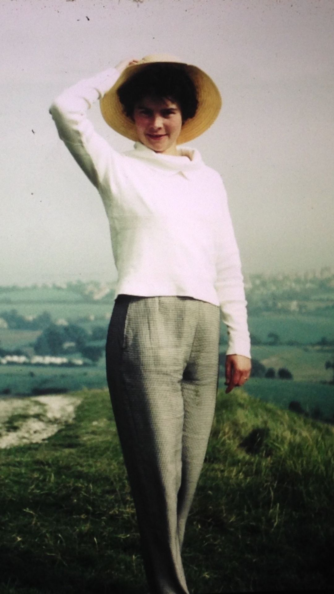 A white woman in a white top and plaid pants stands on a grassy hillside, smiling and holding a straw hat. The background features a lush, misty landscape.