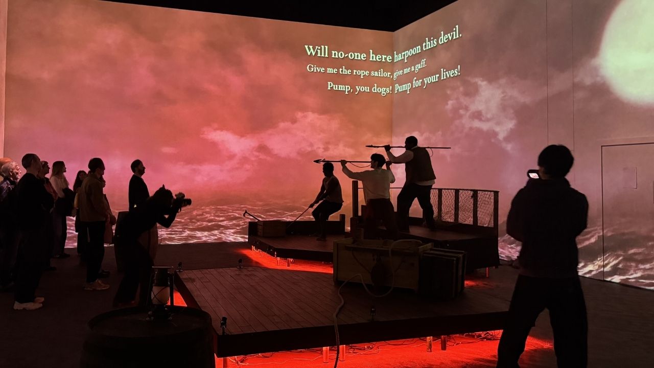 An interactive exhibit with people simulating a harpoon scene against a stormy sea backdrop. Text above reads dramatic maritime dialogue. Atmosphere is intense and immersive.
