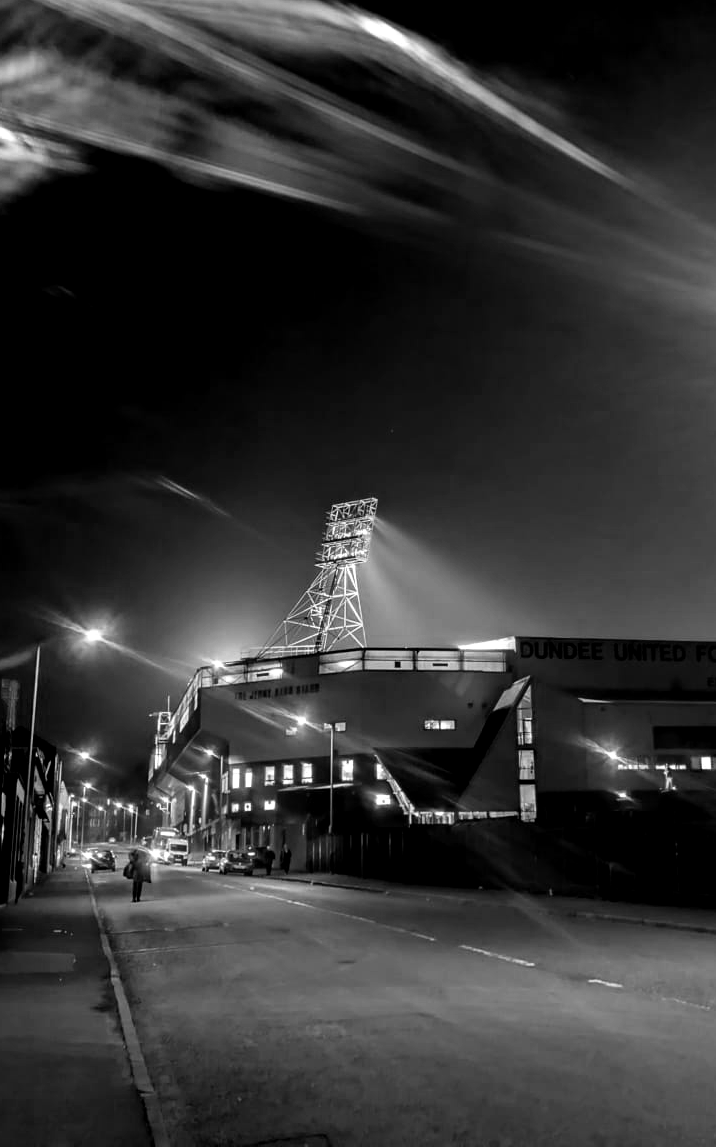 A nighttime street scene features a stadium with a large illuminated tower casting light beams into the sky. A lone person walks on the sidewalk.