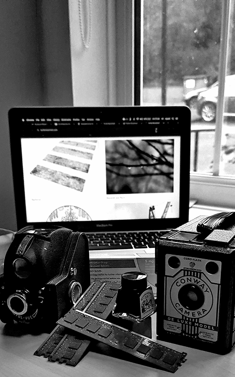 A black-and-white photo of a vintage camera collection on a desk with film negatives, ink bottle, and an open laptop displaying images, near a window.