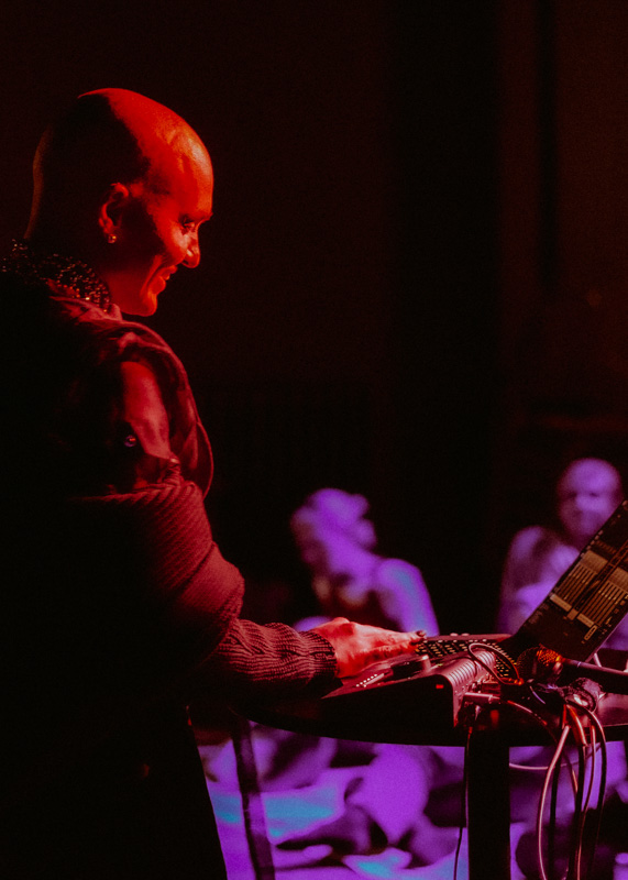 The scene has red and purple lighting. A Brown person with a shaved head smiles softly and is using some music tech. In the background a few blurry people are sat down.