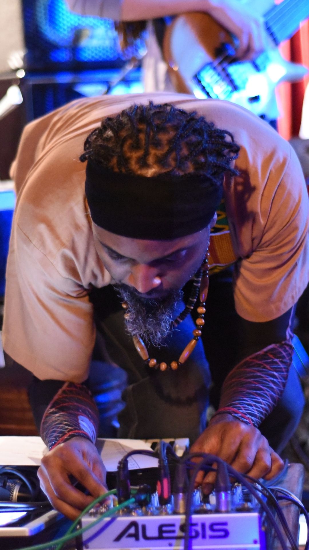 A Black musician with braided hair adjusts audio equipment during a live performance. He's surrounded by instruments, with a focused and creative atmosphere.