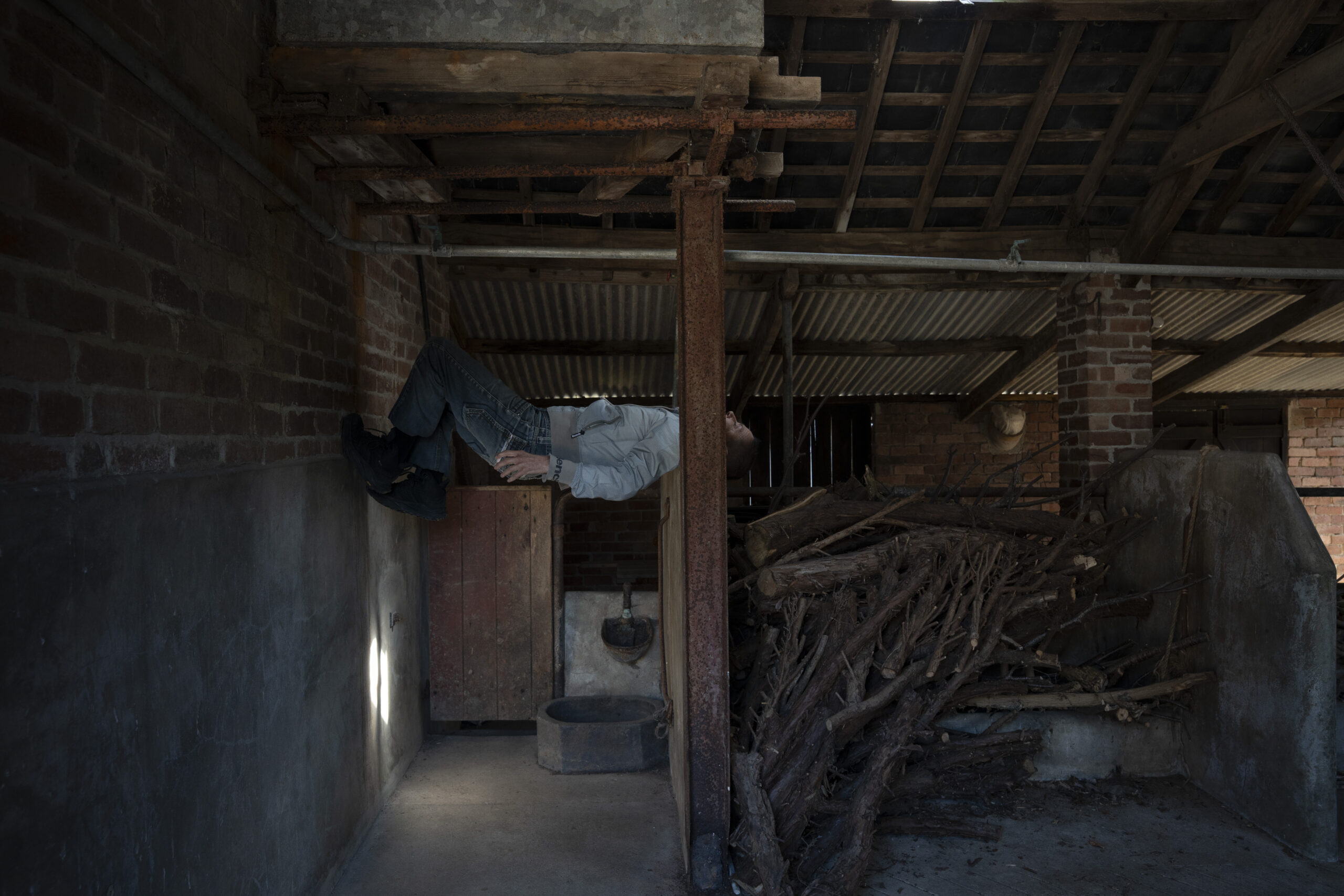 A person lies horizontally across rustic beams in an old barn, creating an illusion of floating. The setting is dimly lit, with wood, bricks, and shadows, giving a mysterious and surreal tone.