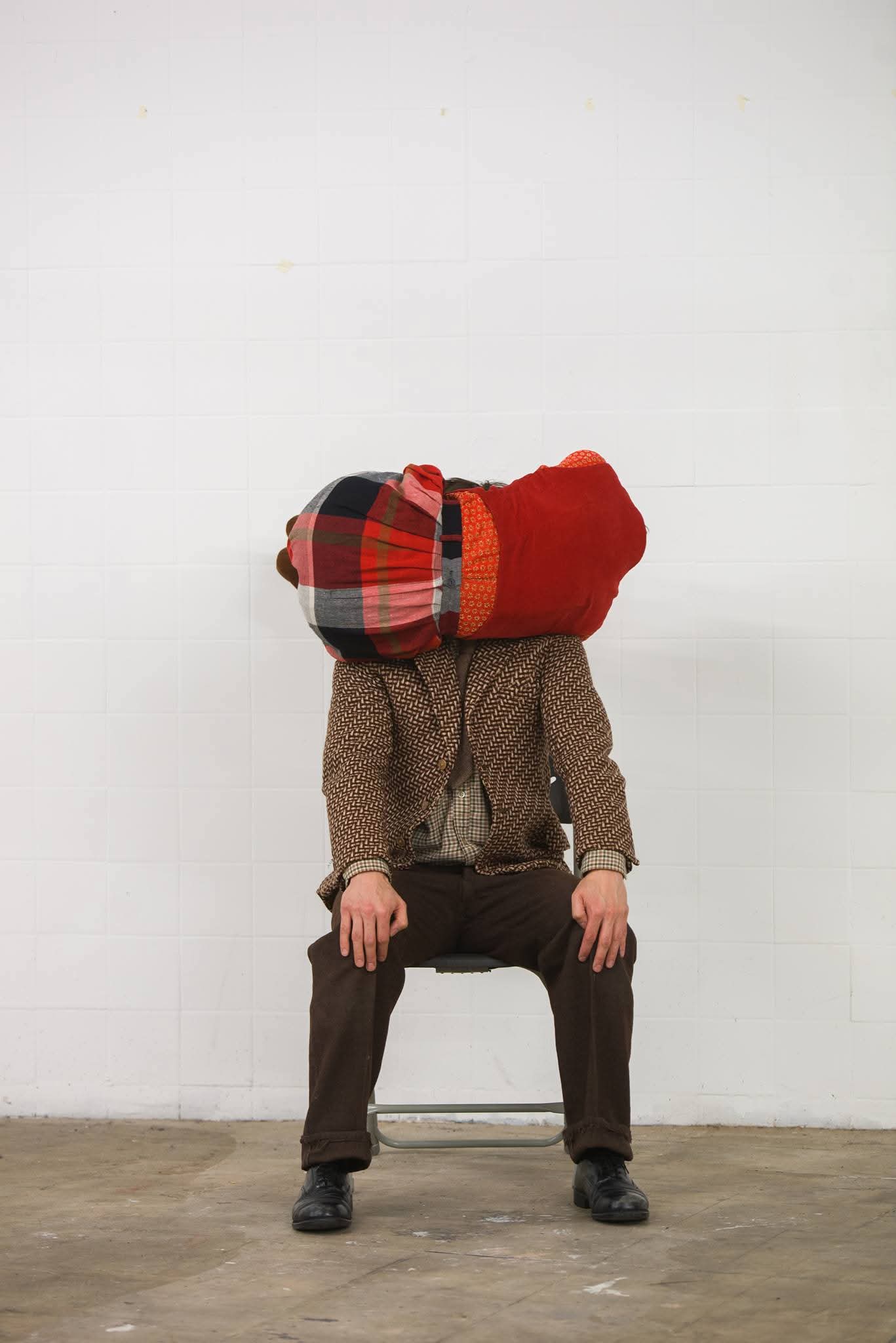 A person sits slumped on a chair in a minimalist room, wearing a brown suit with a large, colorful plaid cushion obscuring their head. The scene conveys a surreal and quirky tone.