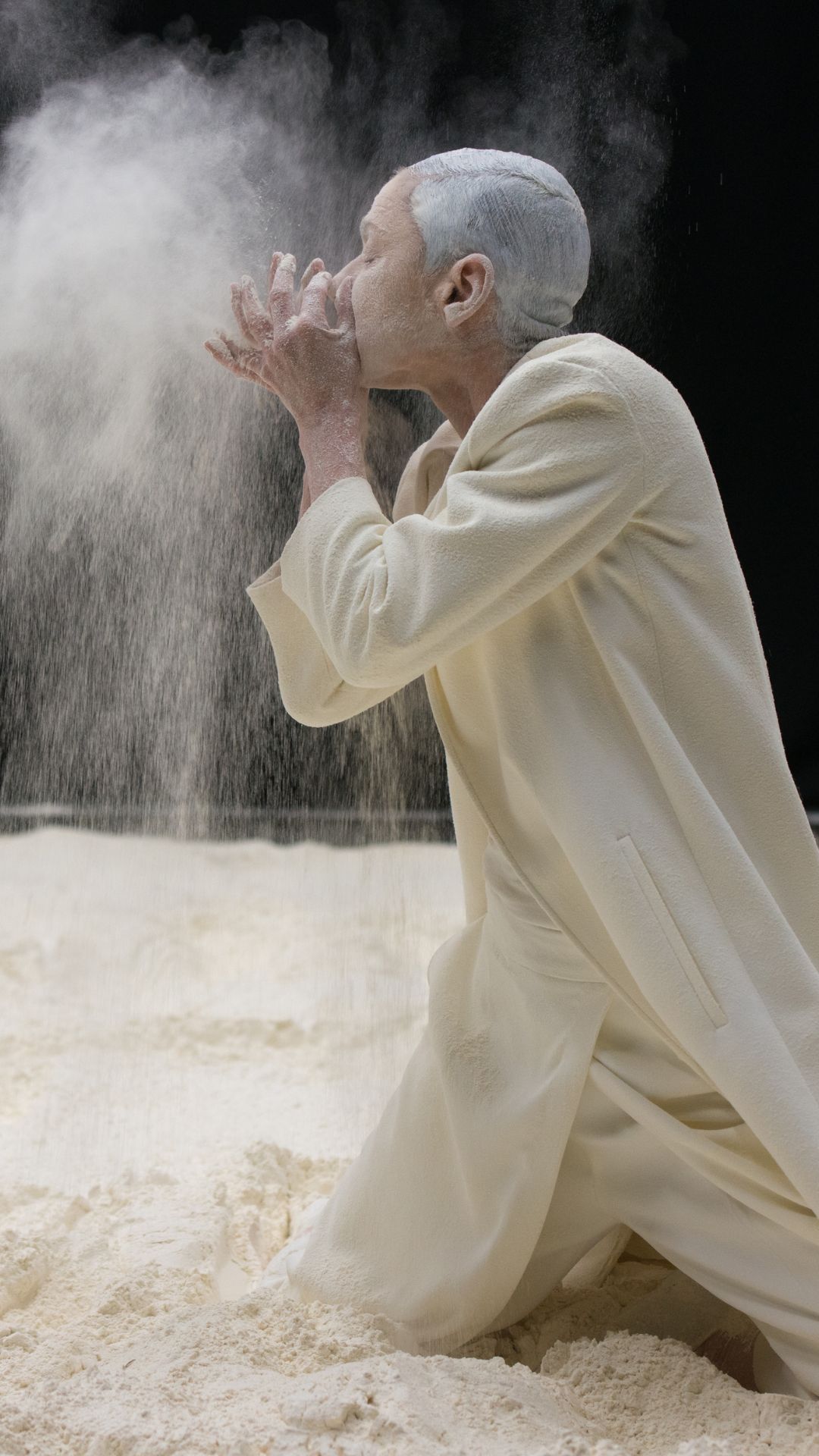 Mae person yn penlinio ar dywod, yn gwisgo gwyn i gyd, yn chwythu cwmwl o bowdr gwyn i'r awyr yn erbyn cefndir tywyll, gan ddeffro naws swrrealaidd, fyfyriol.