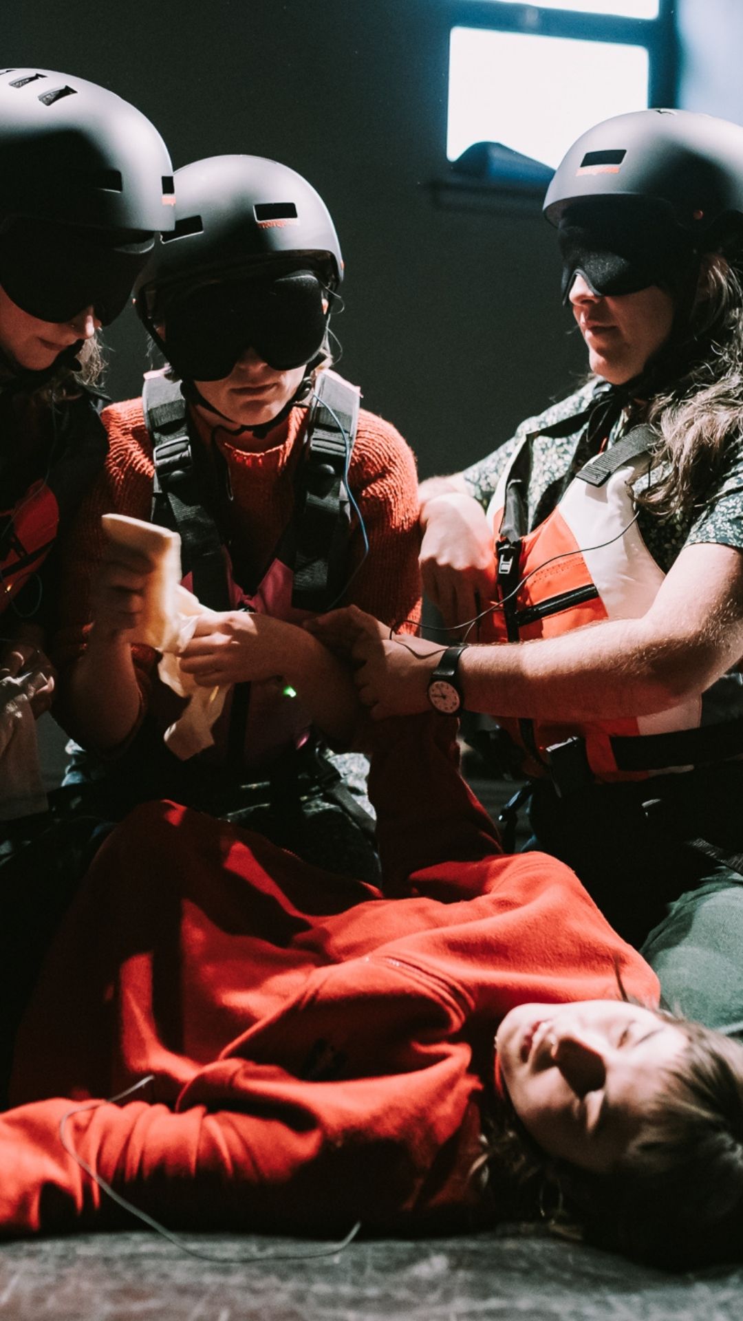 Four people wearing helmets and life vests assist a person lying on the floor in a dramatic scene. The lighting is dim, creating a tense atmosphere.