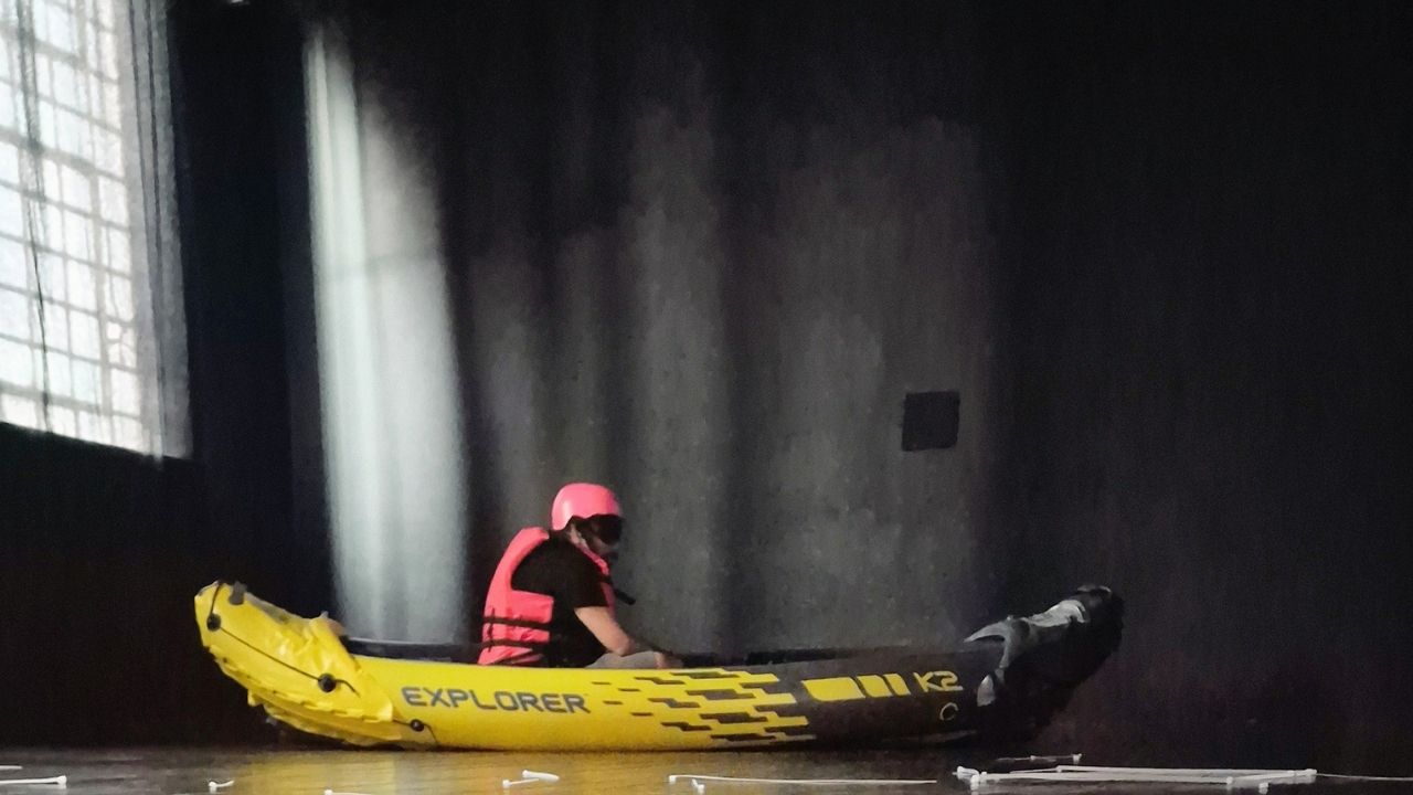Person in a yellow inflatable kayak and pink helmet sits in a dark room with a single window. The lighting casts a dramatic, isolated atmosphere.