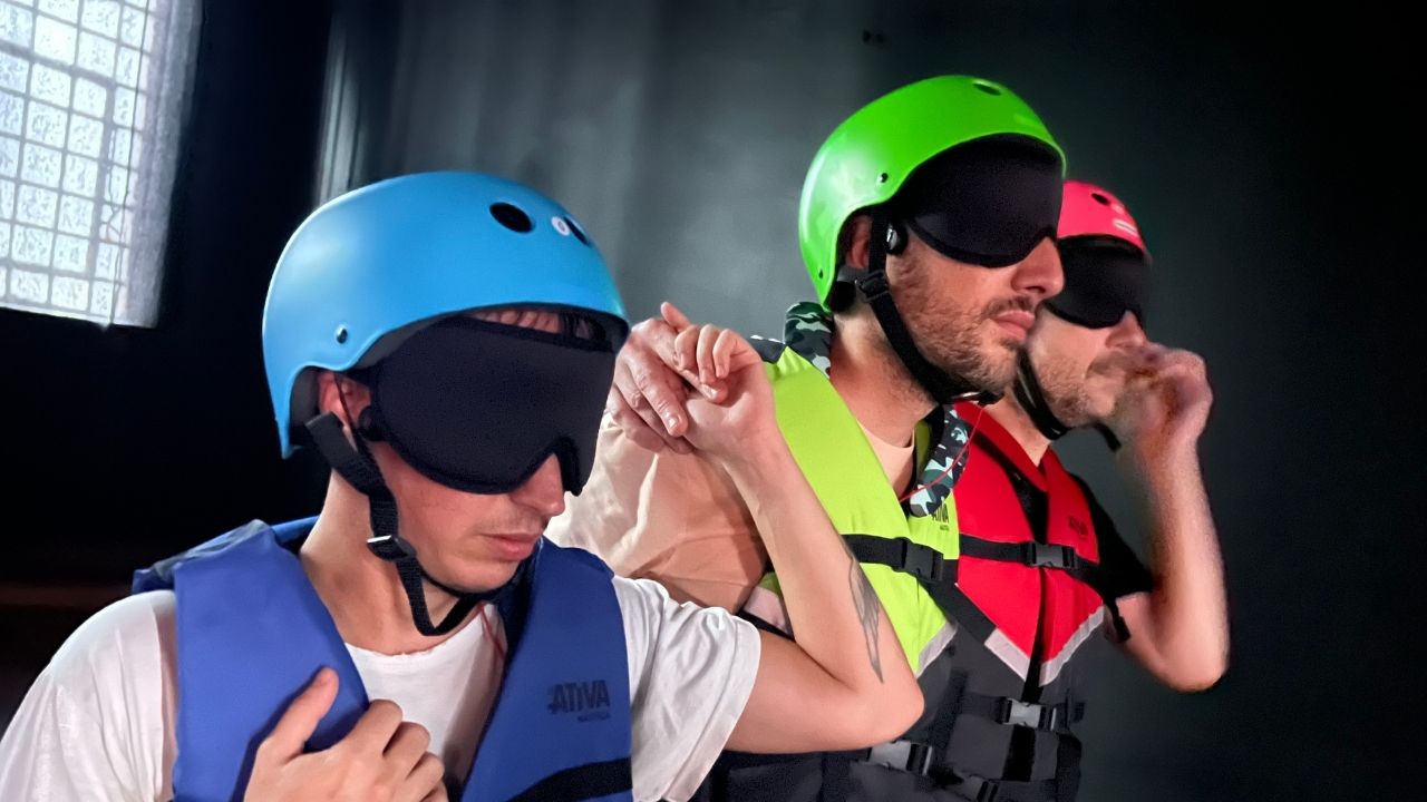 Three people in helmets and life vests, blindfolded, move in line holding shoulders. The scene conveys trust and teamwork in a dimly lit setting.