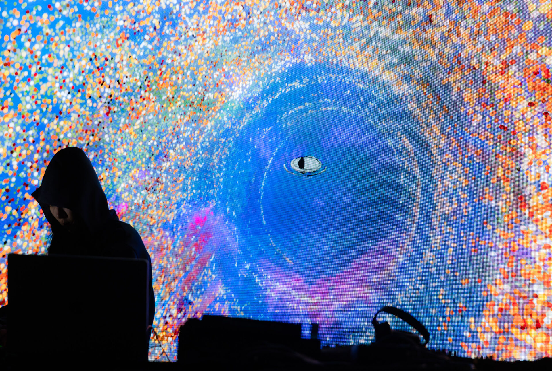 A hooded figure and equipment silhouette against a vivid digital installation, where a blue eye-like orb swirls amid colorful pixel-like lights, creating a surreal mood.