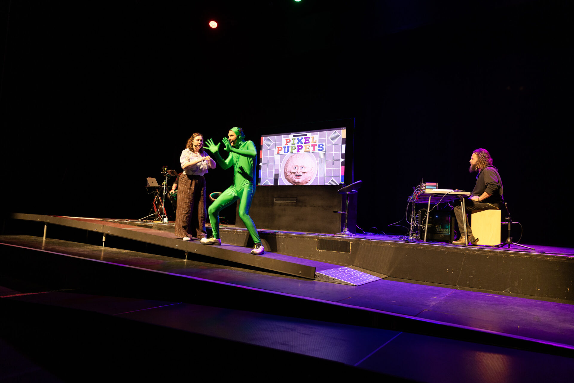 On a dim theatre stage, a performer in a bright green suit interacts with a woman near a screen reading “Pixel Puppets,” as a musician plays keyboard.